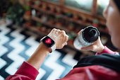 A close-up of a person holding a water bottle while checking their heart rate on a fitness tracker worn on their wrist. The fitness tracker display shows a heart icon, indicating active heart rate monitoring. The background features a zigzag-patterned floor, adding a dynamic focus to the image.